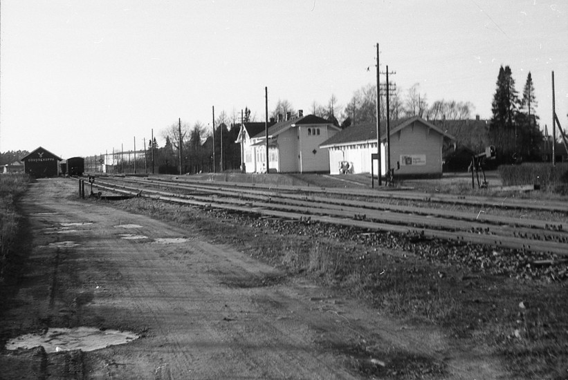 Mustavalkoinen kuva Piikkiön puurakenteisesta rautatieasemasta vieressä kuoppainen ja märkä hiekkatie